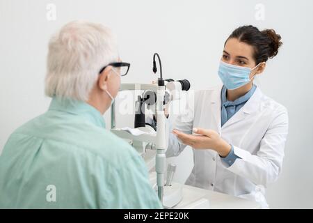 Junge Brünette weibliche Augenarzt in whitecoat und Schutzmaske gehen Um das Augenlicht eines älteren männlichen Patienten zu testen, der davor sitzt Von ihr Stockfoto