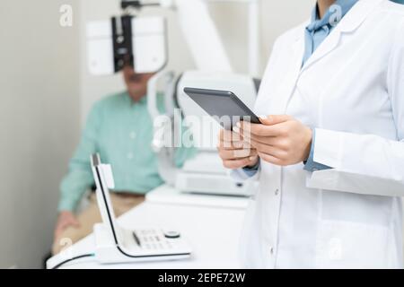 Hände von jungen weiblichen Augenarzt in Whitecoat mit digitalen Tablette Vor dem Hintergrund eines älteren männlichen Patienten, der sein Sehvermögen überprüft hat Nach oben Stockfoto