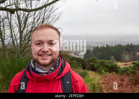 Männlicher Wanderer im Regen lächelt in einer roten Jacke, mit einem Rucksack, Gesichtshaar vor einem Tal bei schlechten Wetterbedingungen in den Highlands. Stockfoto