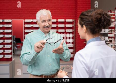 Glücklicher älterer Mann, der jungen Optikerinnen seine Brille zeigt In Whitecoat während der Beratung mit ihr über neue Brillen in Kliniken Stockfoto