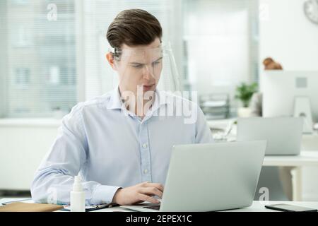 Junge seriöse männliche Manager mit Schutzschirm auf Kopf suchen Auf dem Laptop-Display, während die Netzwerkumgebung vom Schreibtisch aus mit der Büroumgebung verglichen wird Stockfoto