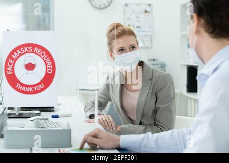 Junge Geschäftsfrau in Schutzmaske Blick auf ihren männlichen Kollegen Sitzen neben ihr während der Diskussion der Arbeitsmomente in Büro Stockfoto