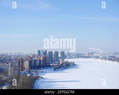 Wolkenkratzer am Ufer des Flusses weiß vom Schnee, in der Ferne am Horizont eine große Stadt Draufsicht Stockfoto