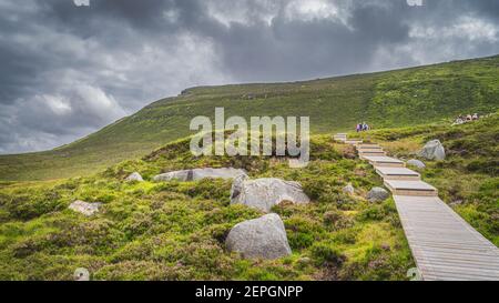 Menschen wandern auf Holzboardwalk, zwischen Felsbrocken und Heiden, die zum Cuilcagh Mountain führen. Sturm, dramatischer Himmel im Hintergrund, Nordirland Stockfoto