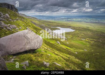 Große Felsbrocken und Schutt am Cuilcagh Mountain Berghang, kleiner See im Tal unten und dramatischer, stürmischer Himmel im Hintergrund, Nordirland Stockfoto