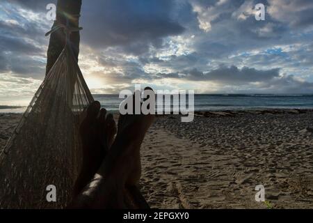 Nahaufnahme der Füße eines Touristen, der sich entspannt, während er das Meer betrachtet, in einer Hängematte an einem tropischen mexikanischen Strand bei Sonnenaufgang. Im Hintergrund der wolkige Himmel. Stockfoto