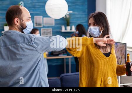 Glückliche fröhliche Freunde berühren Ellbogen halten soziale Distanzierung Gruß eachother tragen Gesichtsmaske verhindern Ausbreitung von Coronavirus hält Bierflaschen in der Wohnung Wohnzimmer. Konzeptuelles Image. Stockfoto