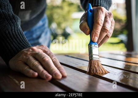 Älterer Mann malt Holztisch. Renovierung von Gartenmöbeln. Nahaufnahme der Hand mit dem Pinsel. Handwerker restauriert Möbel im Freien Stockfoto