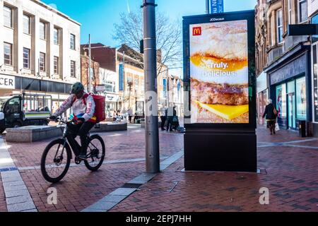 Ein gerade essen Lieferung Radfahrer fährt vorbei an einer elektronischen Werbung JCDecaux Plakatwand in Broad Street, Reading, Großbritannien, die eine McDonalds Anzeige zeigt. Stockfoto