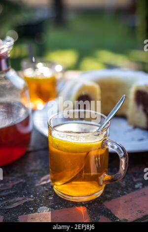Erfrischung mit Tee und gebundenem Kuchen im Garten. Frühstück am Morgen im Freien. Glas Kräutertee mit Zitrone und Löffel auf dem Tisch Stockfoto