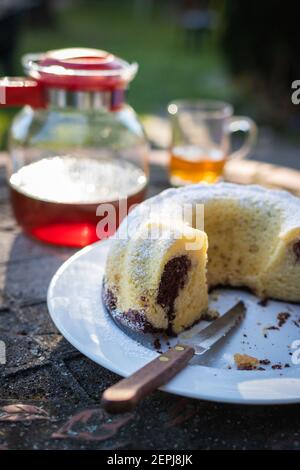 Erfrischung mit gebundenem Kuchen und Tee im Garten. Frühstück am Morgen im Freien. Biskuitkuchen und Messer auf weißem Teller am Tisch Stockfoto