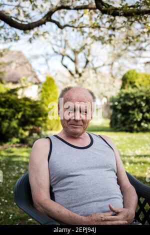 Porträt eines glücklichen älteren Mannes, der in seinem Garten auf einem Stuhl sitzt. Alter Mann mit lächelndem Gesicht, der im Frühling im Hinterhof relaxt. Blick auf die Kamera Stockfoto