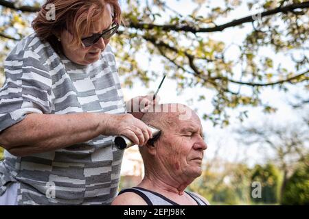 Ältere Frau macht Haarschnitt mit Haarschneider zu ihrem Mann im Garten. Ältere Menschen kümmern sich gegenseitig. Häusliches Leben Stockfoto