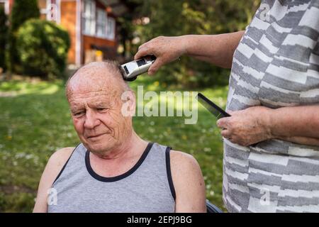 Frau macht Frisur mit Haarschneider zu älteren Mann im Freien im Garten. Ältere Menschen kümmern sich während der Pandemiesperre um einander Stockfoto