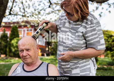 Frau macht Haarschnitt mit Haarschneider zu älteren Mann im Freien im Garten. Ältere Menschen kümmern sich gegenseitig. Glückliches älteres Paar Stockfoto