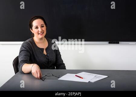 Eine junge Lehrerin sitzt an ihrem Schreibtisch vor einer Tafel. Stockfoto