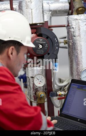 Zentralheizung. Thermometer an der Warmwasserzuleitung zum Heizsystem. Techniker trägt rote Jacke mit weißem Helm. Stockfoto