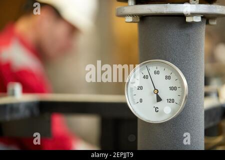 Zentralheizung. Thermometer an der Warmwasserzuleitung zum Heizsystem. Techniker trägt rote Jacke mit weißem Helm. Stockfoto