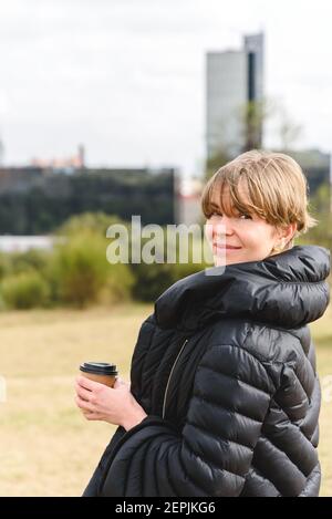 Vertikales Porträt einer blonden kaukasischen jungen Frau, die stilvolles schwarzes Winterkleid kleidet, eine Einweg-Tasse Kaffee hält und die Kamera mit Stadtgebäuden im Hintergrund anschaut Stockfoto
