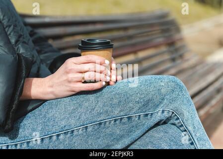 Nahaufnahme der Hände einer weißen Frau, die eine Einweg-Papiertasse mit Kaffee hält. Sie trägt blaue Jeans, schwarzen Mantel und einen Ring an den Fingern. Sie sitzt auf einer Holzbank in einem städtischen grünen Park Stockfoto