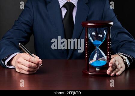 Geschäftsmann hält Stift an seinem Arm und Sanduhr auf einem Tisch stehen. Deadline Konzept Stockfoto