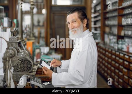 Angenehmer älterer männlicher Apotheker, der an digitalen Tabletten arbeitet. Regal mit alten Medikamentenschachteln und altem Kassenregal im Hintergrund. Medizin Stockfoto