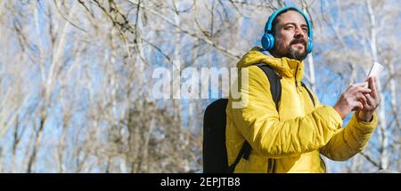 Junger Hipster-Mann mit Bart und Rucksack Zurück Musik hören mit seinen Kopfhörern und seinem Handy Telefon in einem Park Stockfoto