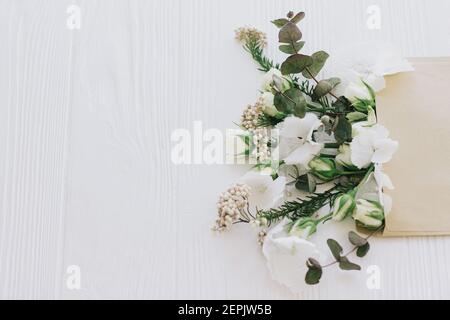 Stilvolle Rosen, Hortensien und Eukalyptuszweige wachsen aus handgefertigte Umschlag auf weißem Holz mit Kopieplatz. Kreatives Frühlingsbild, florale Begrüßung ca. Stockfoto