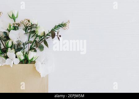 Stilvolle Rosen, Hortensien und Eukalyptuszweige wachsen aus Handwerk Umschlag auf weißem Holz, flach legen mit Kopie Raum. Blumen Grußkarte und Blume Stockfoto