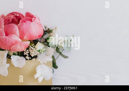 Pfingstrose, weiße Blumen und Eukalyptus wachsen aus Bastelhülle auf weißem Holz, flache Laie mit Platz für Text. Glücklicher Muttertag und Frauentag. Creativ Stockfoto