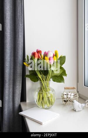 Ein Bukett aus bunten Tulpen in einer transparenten Vase auf der Fensterbank im Urlaub. Weißes Notizbuch, Korbkorb aus Metall mit Baumwollblumen innen Stockfoto