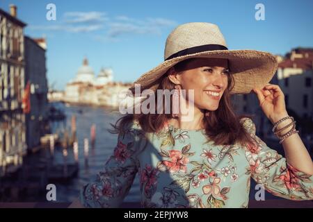 Lächelnd elegante Solo-Reisende Frau in Blumenkleid mit Hut mit Rundgang auf Accademia Brücke in Venedig, Italien. Stockfoto