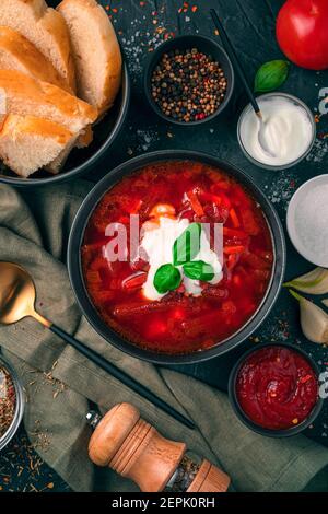 Ein Teller Rote Beete Suppe mit saurer Sahne und Rosmarin, Brot und Tomatensauce auf schwarzem Beton Hintergrund. Stockfoto