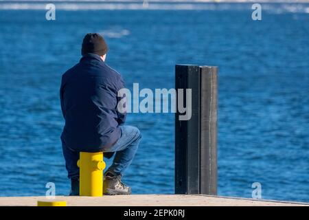 Helsinki / Finnland - 27. FEBRUAR 2021: Ein alleinstehender Mann sitzt auf einem gelben Poller und blickt auf das gefrorene Meer Stockfoto