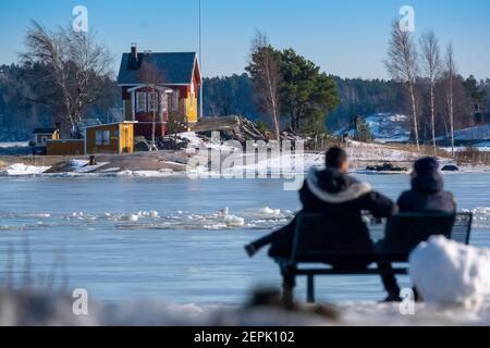 Helsinki / Finnland - 27. FEBRUAR 2021: Zwei Männer sitzen auf einem Strandboulevard und blicken auf ein altes Holzhaus auf der Insel. Stockfoto