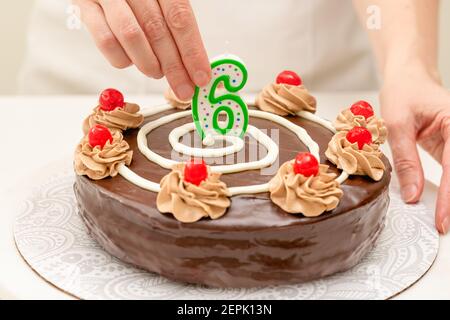 Geburtstagskuchen mit Schokolade. Frau Hände Dekoration Kuchen mit Kerze in Form von Nummer sechs. Hausgemachter Kuchen mit Schokoladencreme und Kirschen dekoriert Stockfoto