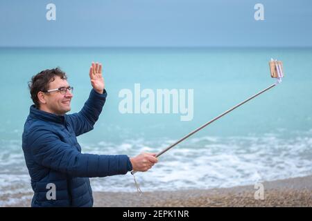 Junger Mann am Strand mit einem Smartphone auf einem Selfie-Stick. Blogger kommuniziert mit Abonnenten. Stockfoto