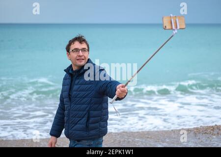 Der junge Mann am Meer fotografiert sich auf einem Smartphone mit einem Selfie-Stick. Stockfoto