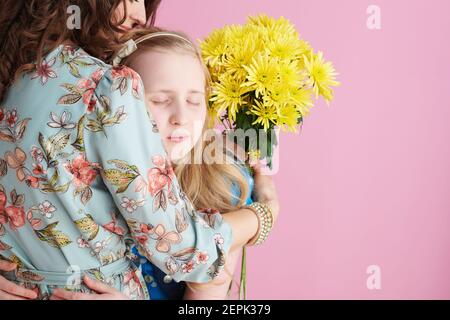 Sentimental trendy Mutter und Kind mit langen welligen Haar mit gelben Chrysanthemen Blumen umarmen isoliert auf rosa. Stockfoto