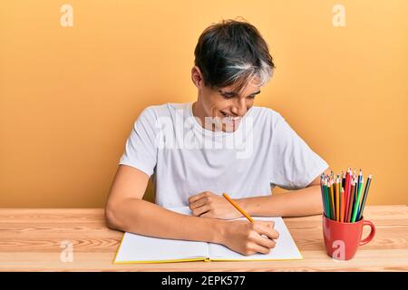 Junger hispanischer Mann schreibt Buch auf dem Tisch sitzen lächelnd und lachen hart aus laut, weil lustig verrückt Witz. Stockfoto