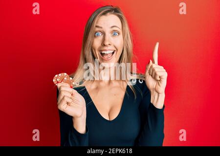 Schöne kaukasische Frau spielt Poker hält Casino-Chips lächeln mit einer Idee oder Frage Zeigefinger mit glücklichen Gesicht, Nummer eins Stockfoto
