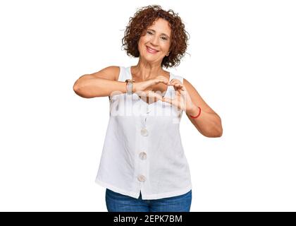 Schöne mittlere Reife Frau tragen lässige weiße Hemd lächelnd in der Liebe tun Herz Symbol Form mit Händen. Romantisches Konzept. Stockfoto