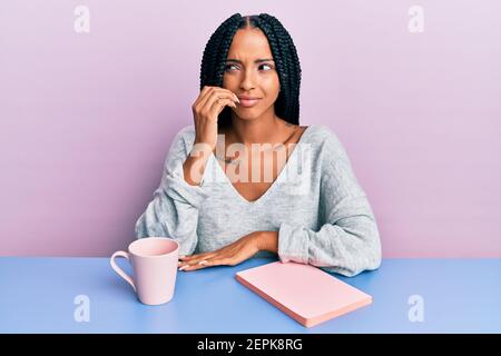 Schöne hispanische Frau, die ein Buch liest und einen Kaffee trinkt, der gestresst und nervös mit den Händen auf den Mund beißt Nägel aussieht. Angstproblem. Stockfoto