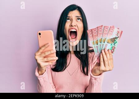 Junge hispanische Frau mit Smartphone hält neuseeländische Dollar Banknoten wütend und verrückt schreien frustriert und wütend, schreien mit Wut schaut Stockfoto