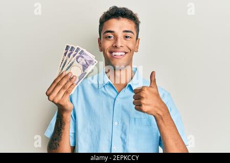 Junger schöner afroamerikanischer Mann hält 5000 japanische Yen Banknoten lächelnd glücklich und positiv, Daumen nach oben tun ausgezeichnet und Zustimmung Zeichen Stockfoto