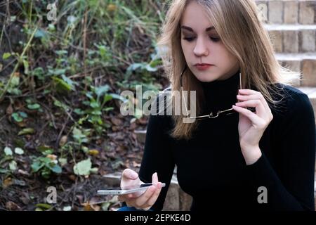 Porträt einer konzentrierten, seriösen Frau, die in ein Mobiltelefon schaut und die Sonnenbrille aus den Augen zieht. Eine Frau sitzt auf den Stufen im Park Stockfoto