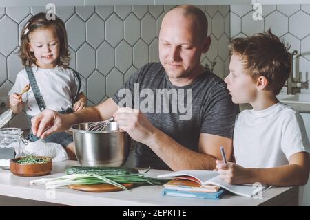 Vater peitscht Omelette mit Schneebesen und hilft seinem Sohn, seine Hausaufgaben mit seiner Tochter neben ihm sitzen. Mann, der Hausaufgaben macht. Stockfoto