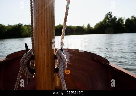 Nahaufnahme der Stollen auf dem Holzmast eines kleinen Segelbootes (Dinghy): Seile (Halyards) in lockeren Knoten gebunden, ein See oder Fluss im Hintergrund Stockfoto