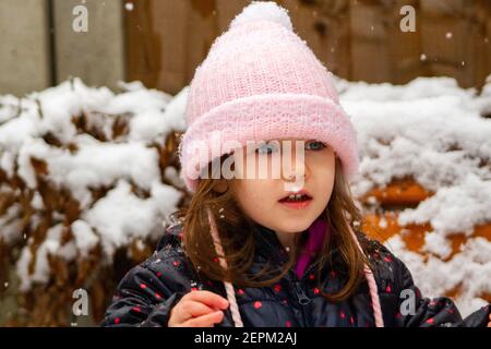 Porträt eines niedlichen, blauäugigen, braunhaarigen Mädchens, das einen rosa Hut und einen ablauen Mantel im Schnee trägt Stockfoto