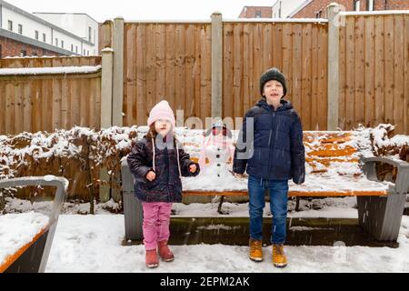 Ein süßer, blauäugiger, rothaariger Junge und ein süßes, blauäugiges, braunhaariges Mädchen, das im Schnee von einem Schneemann steht Stockfoto